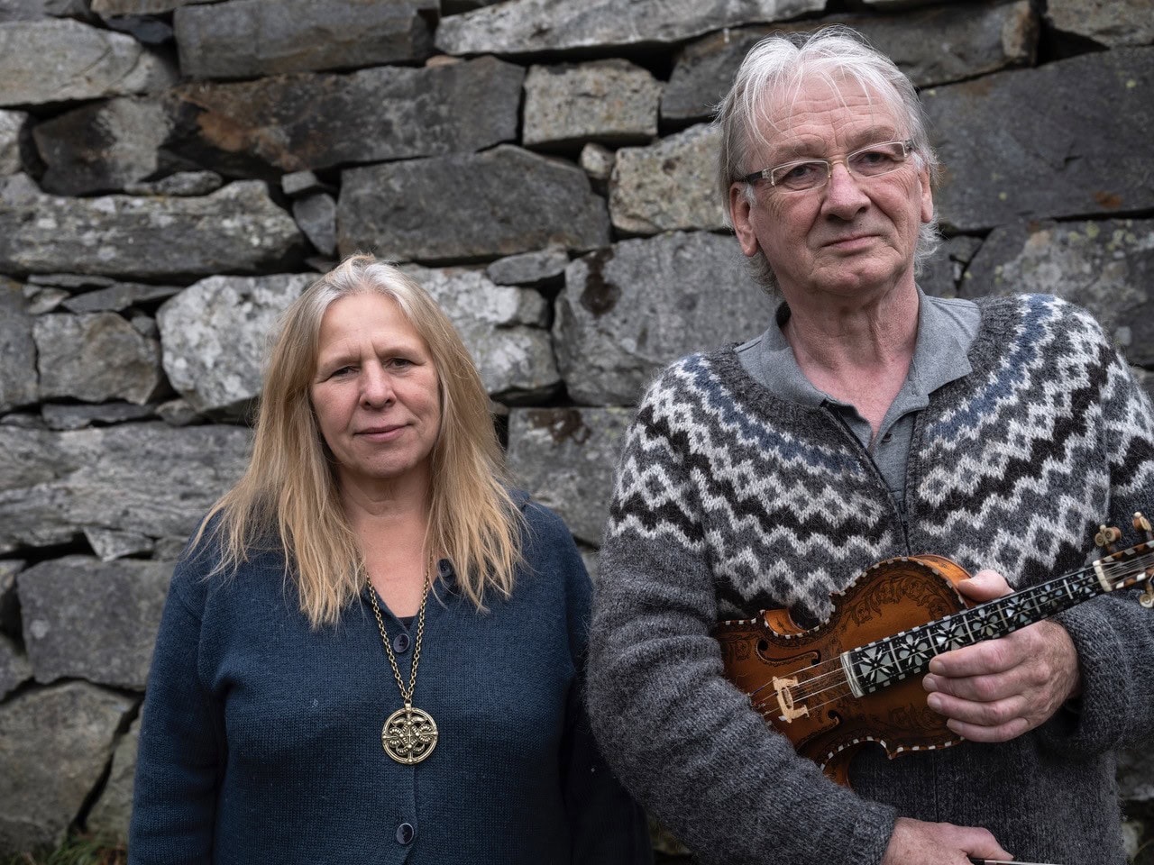 Knut Hamre og Sinikka Langeland framfor ein mur. Knut med fela i handa.